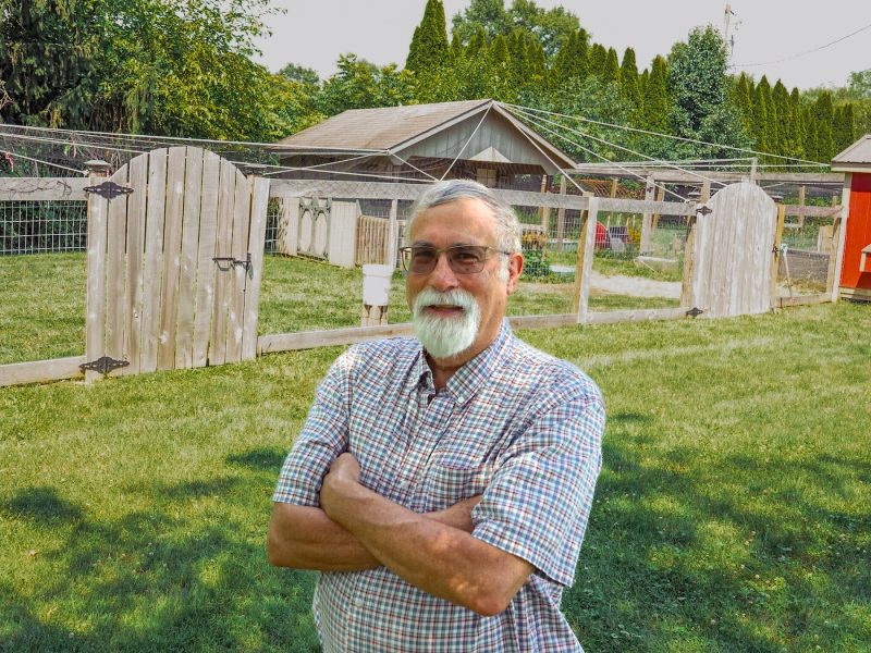 Gary Watson stands in his yard in front of his chicken run, protected from predators by bird netting and wooden fencing.